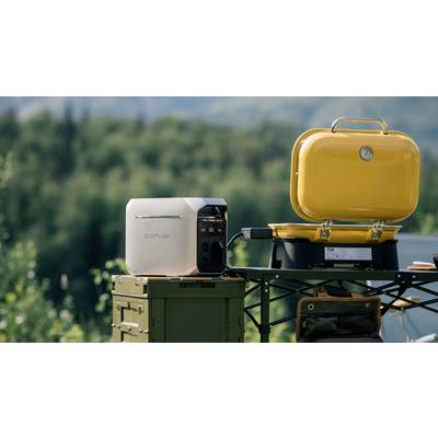 A portable EcoFlow power station sitting on a table next to a yellow barbecue grill outdoors. Trees and mountains are visible in the background.
