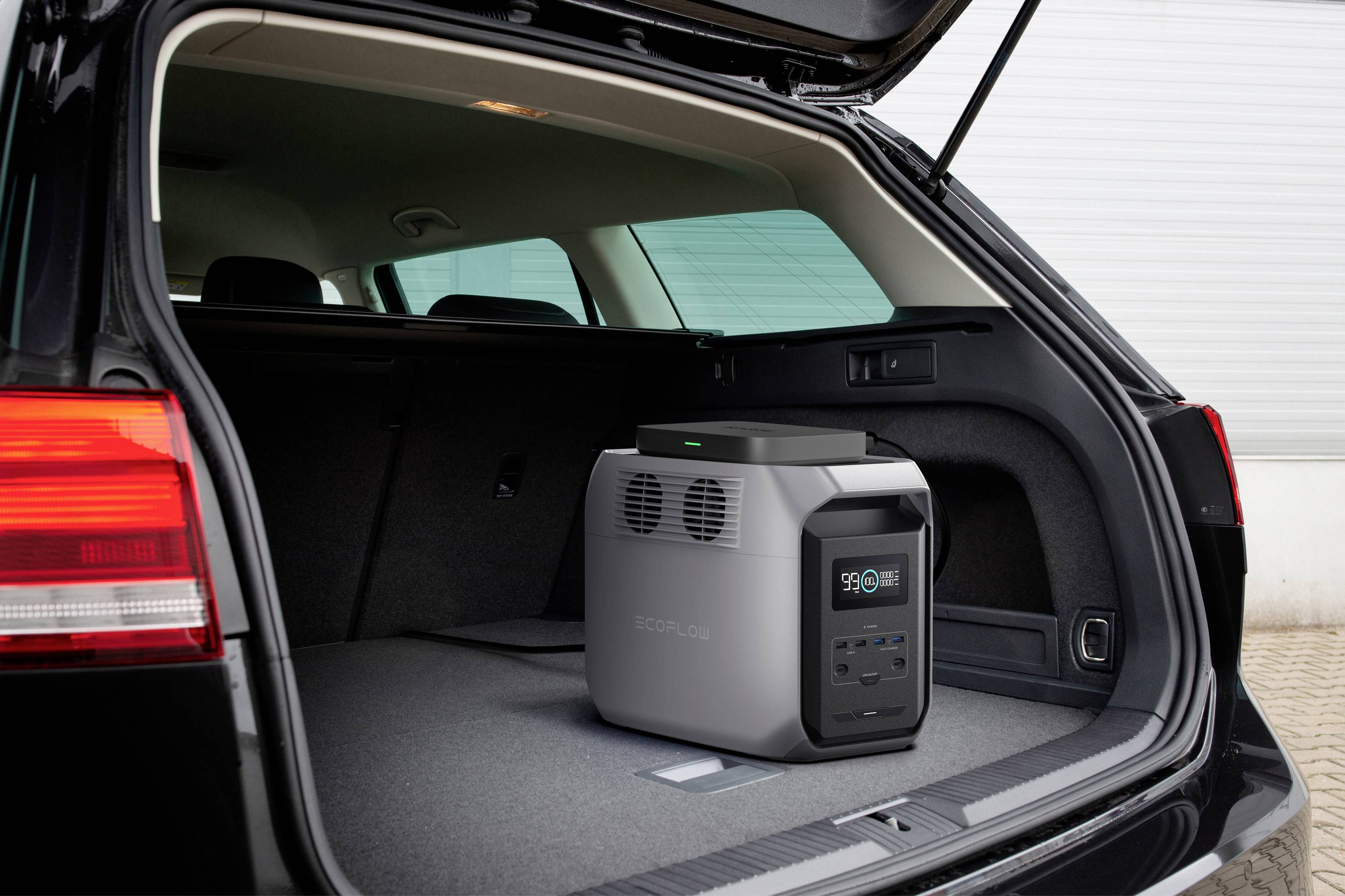 A portable power generator with a digital display showing '90%' is situated in the open boot of a car in front of a white wall.