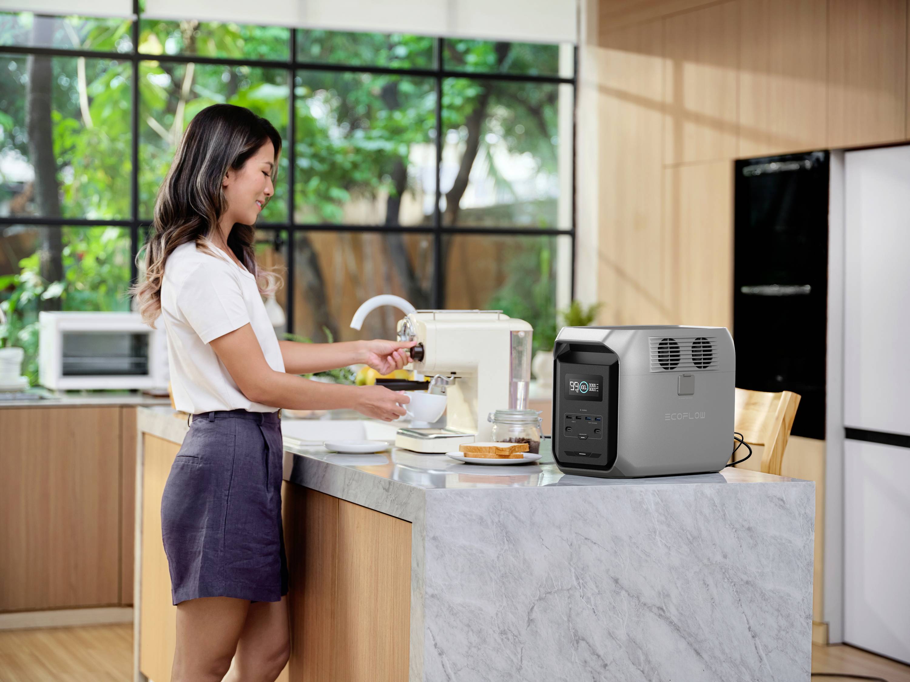 A woman in a modern kitchen is using a food processor. Beside her is a grey battery unit with a display. Windows in the background reveal green trees.