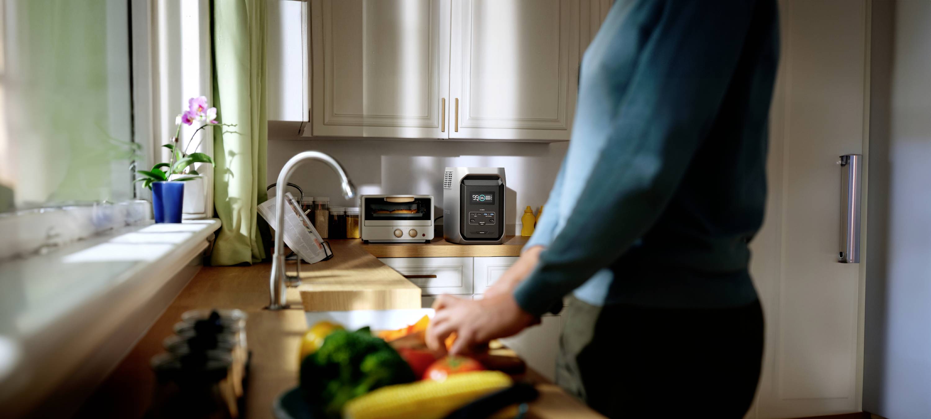 A person is chopping vegetables in a modern kitchen. A toaster and a coffee machine are sitting on the worktop. A window with a flower is in the background.