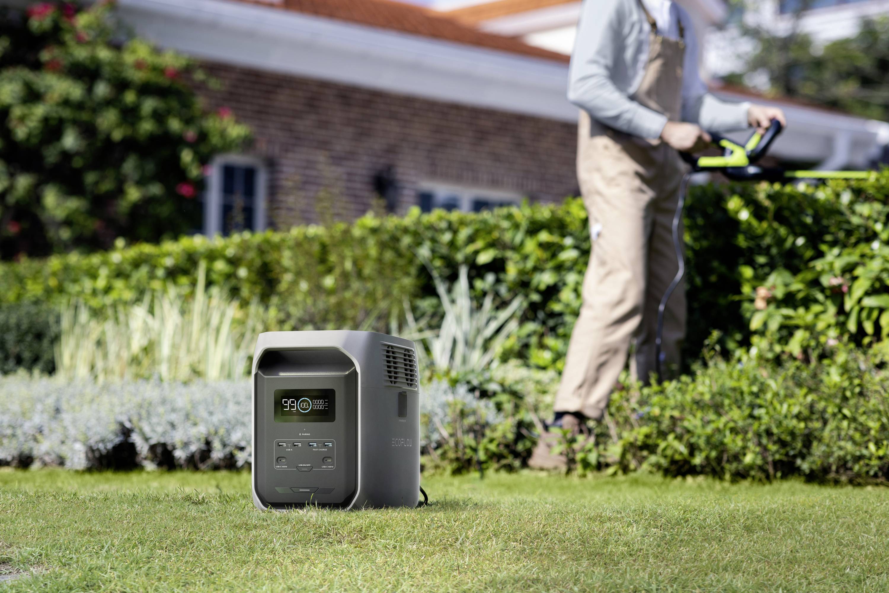 A portable power station sits on a lawn, while a person works in the background with a strimmer.