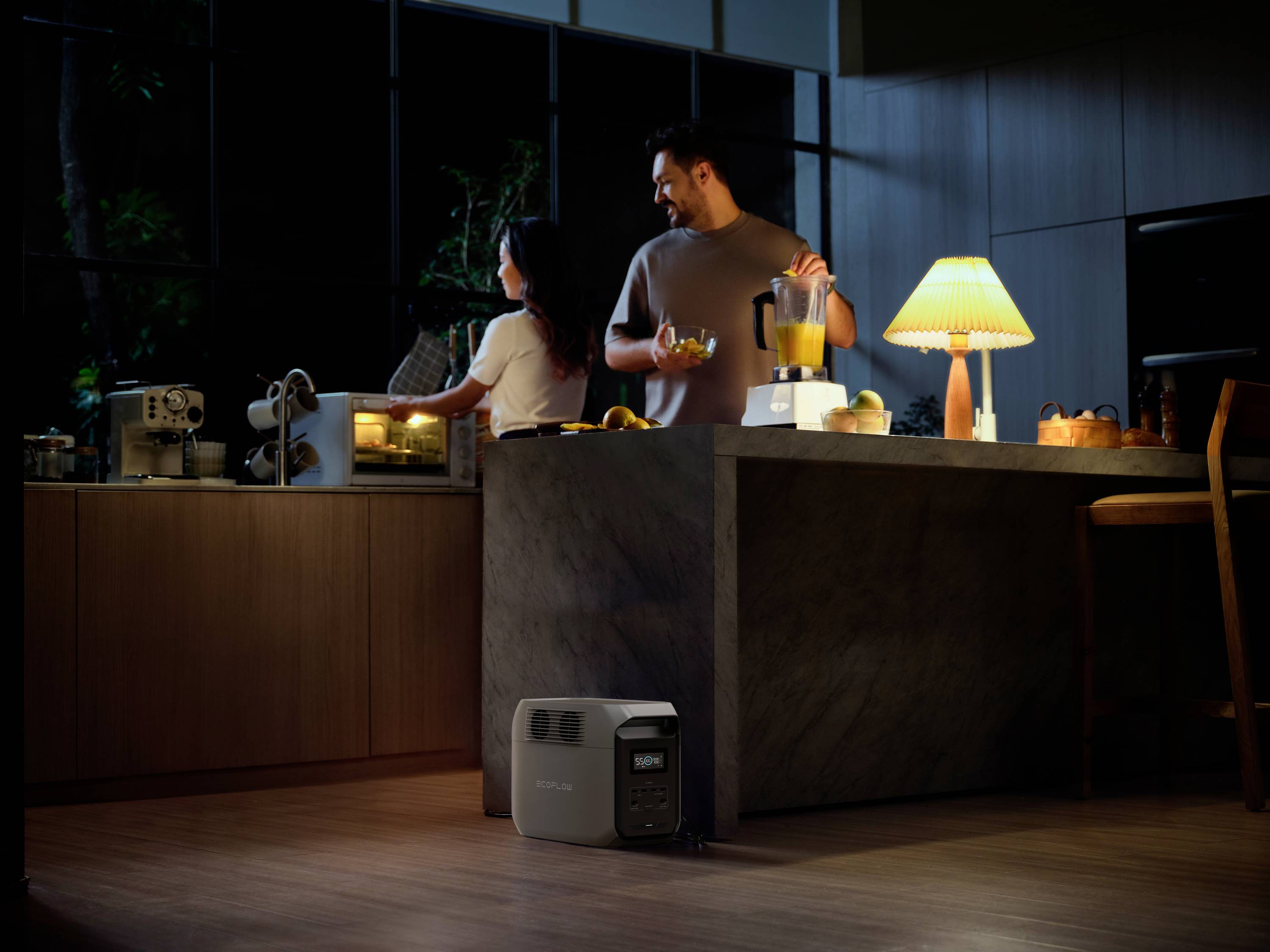 A man and a woman in a modern kitchen. The man is using a blender, the woman is looking into an oven. A lamp illuminates the room.