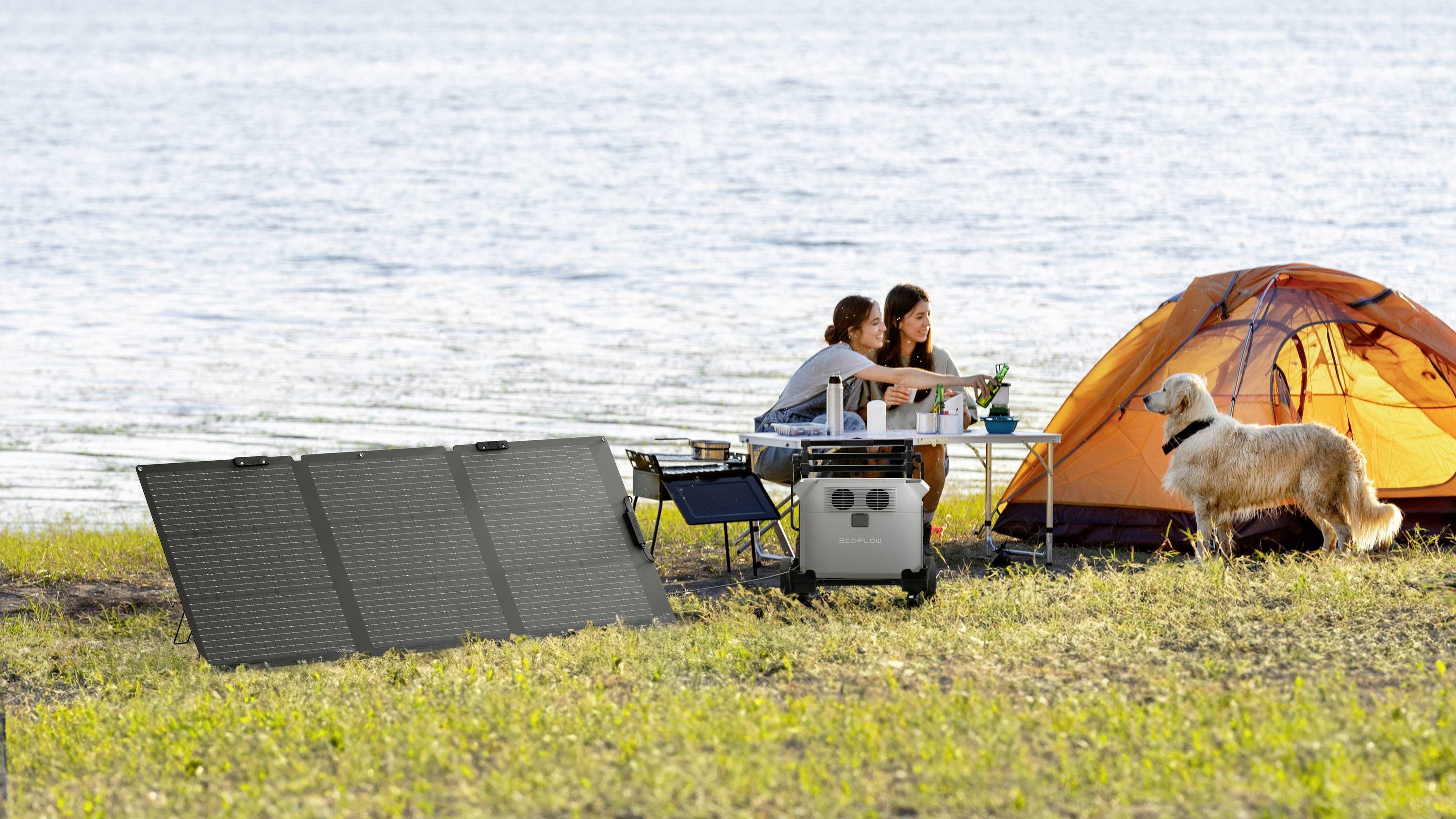 A woman and a man are sitting at a table next to a tent by the lake. They are using a portable solar power system. A dog is standing beside them.