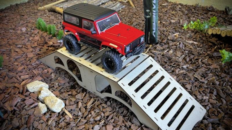 A red remote-controlled car drives over a small metal ramp in a landscape with wood chippings and a plant model.