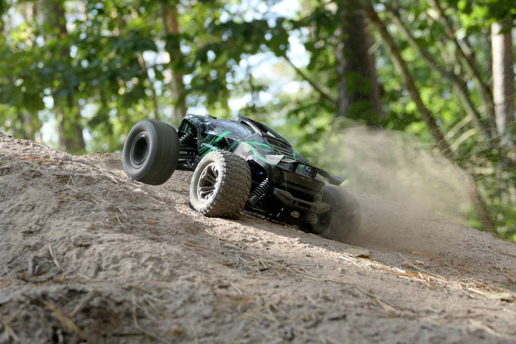 A remote-controlled car speeds up a dusty hill in the woods. Trees and sunlight in the background.