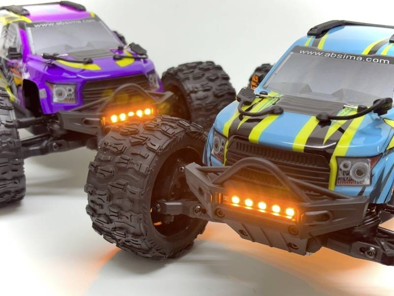 Two remote-controlled trucks in vibrant colours are parked side by side. They are blue and purple, with large tyres and bright headlamps.
