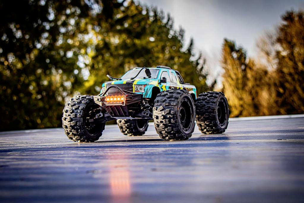 A remote-controlled car with large tyres, driving on a smooth surface outdoors. Blurry trees are visible in the background.