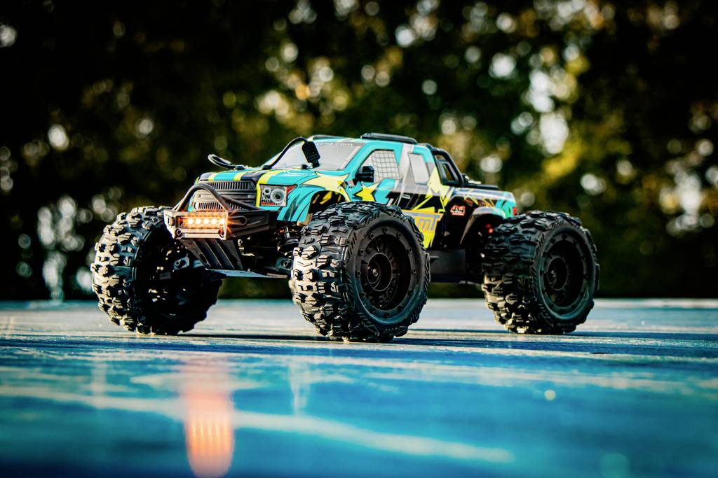 A remote-controlled car with large tyres and a colourful design drives across a smooth surface outdoors, surrounded by trees in the background.