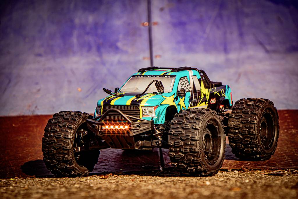 A blue-black remote-controlled car with yellow stars drives on a dirty surface in front of a blue wall.