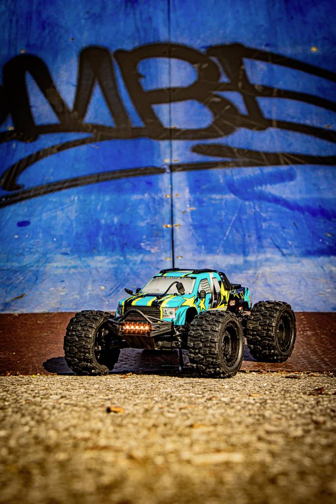 A colourful toy car drives on a rough surface in front of a wall with blue graffiti. The car is off-road capable.