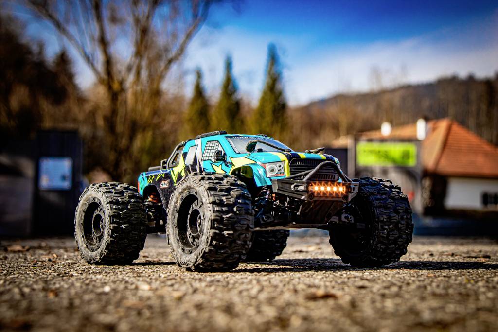 A remote-controlled car with large tyres drives along an unpaved path. Trees and buildings are visible in the background.