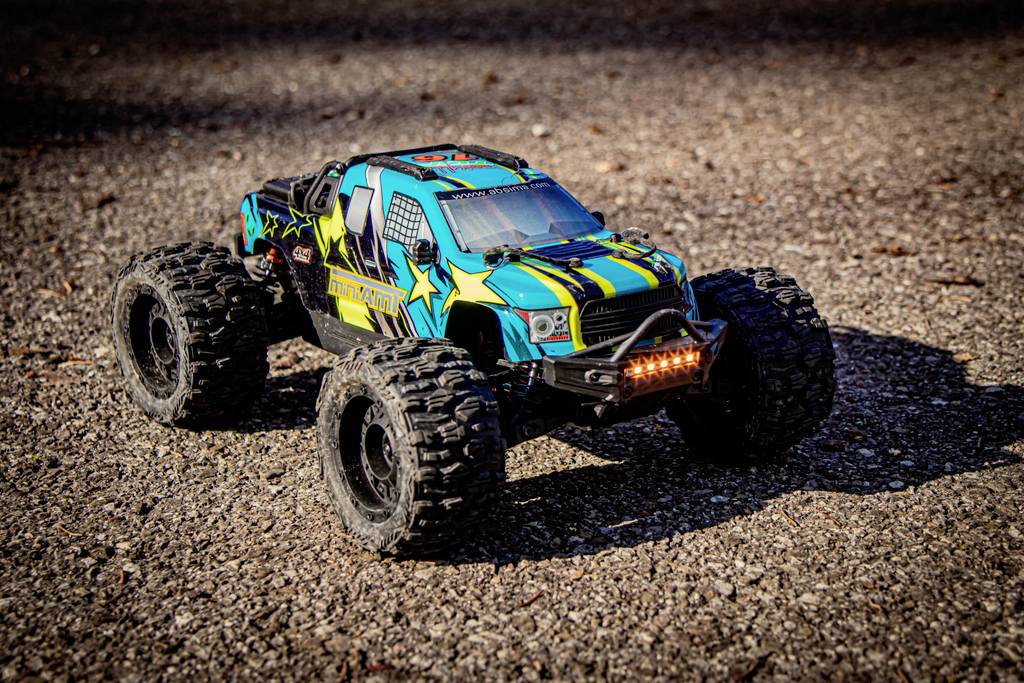 A remote-controlled car with large tyres and colourful patterns sits on a gravel path, illuminated by sunlight.
