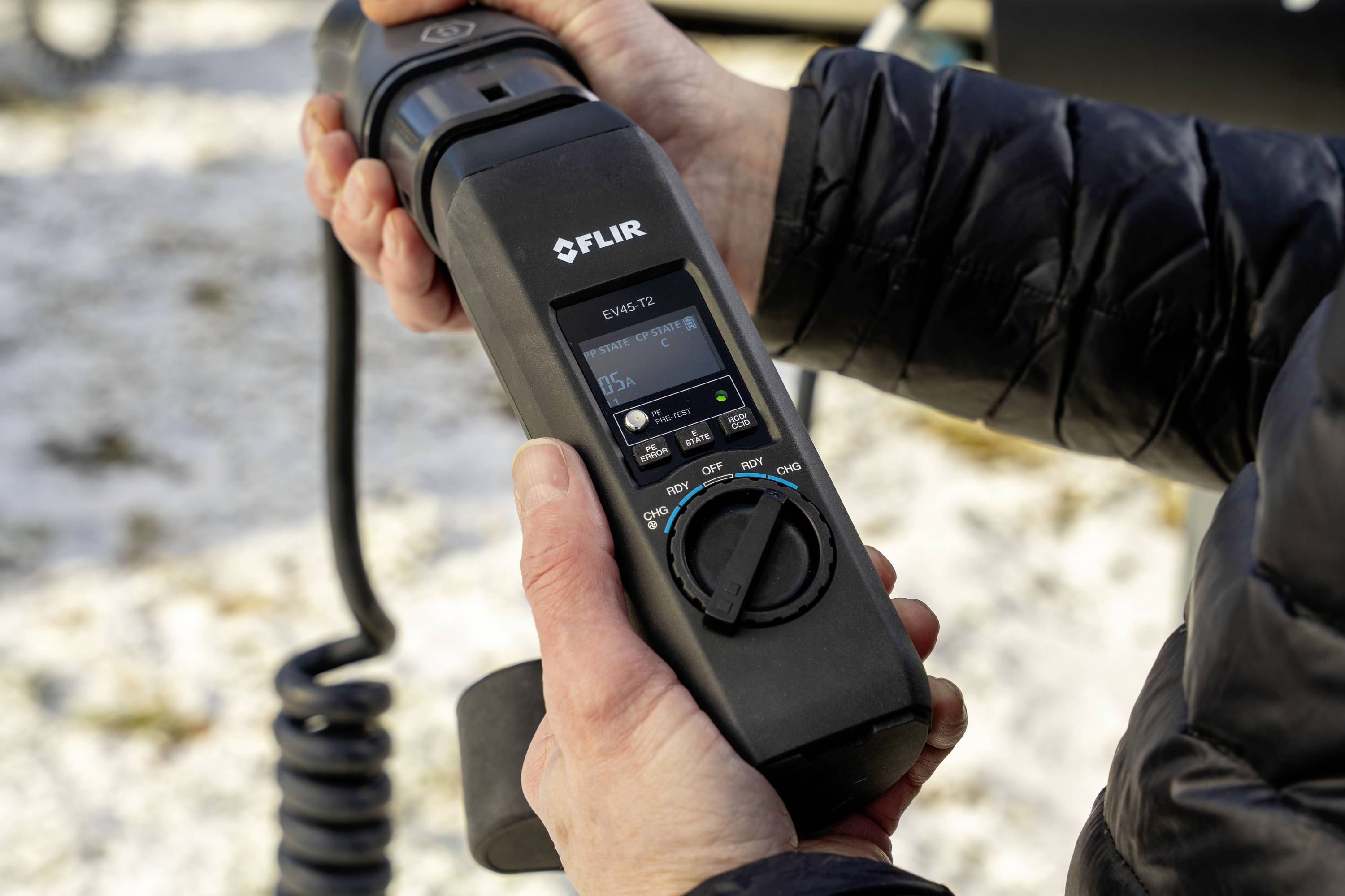 A person is holding a FLIR handheld device with a digital display, used for temperature measurements or diagnostics outdoors.