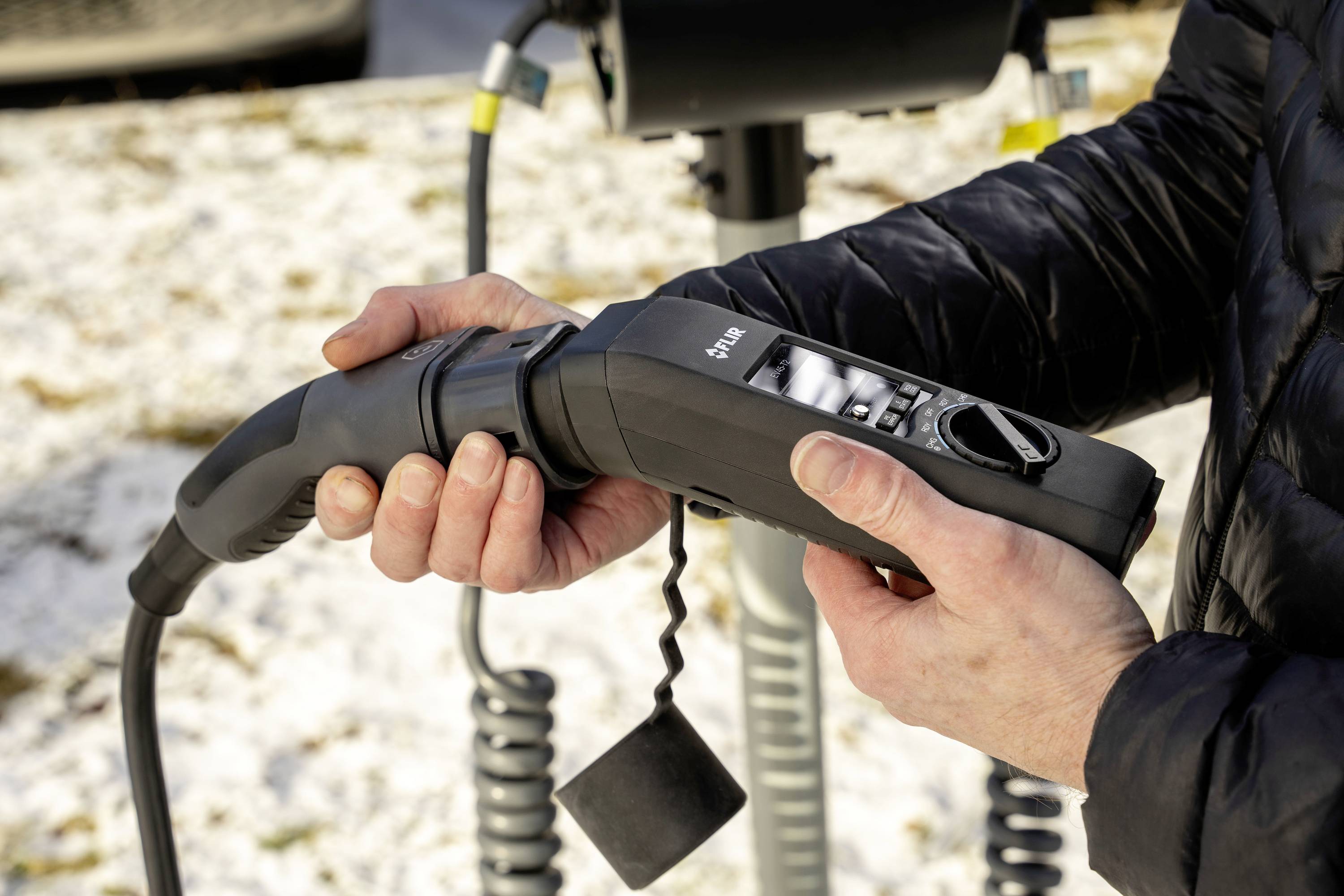 A person is holding an electric vehicle charging cable in their hands and connecting it to a vehicle. The background shows snow.