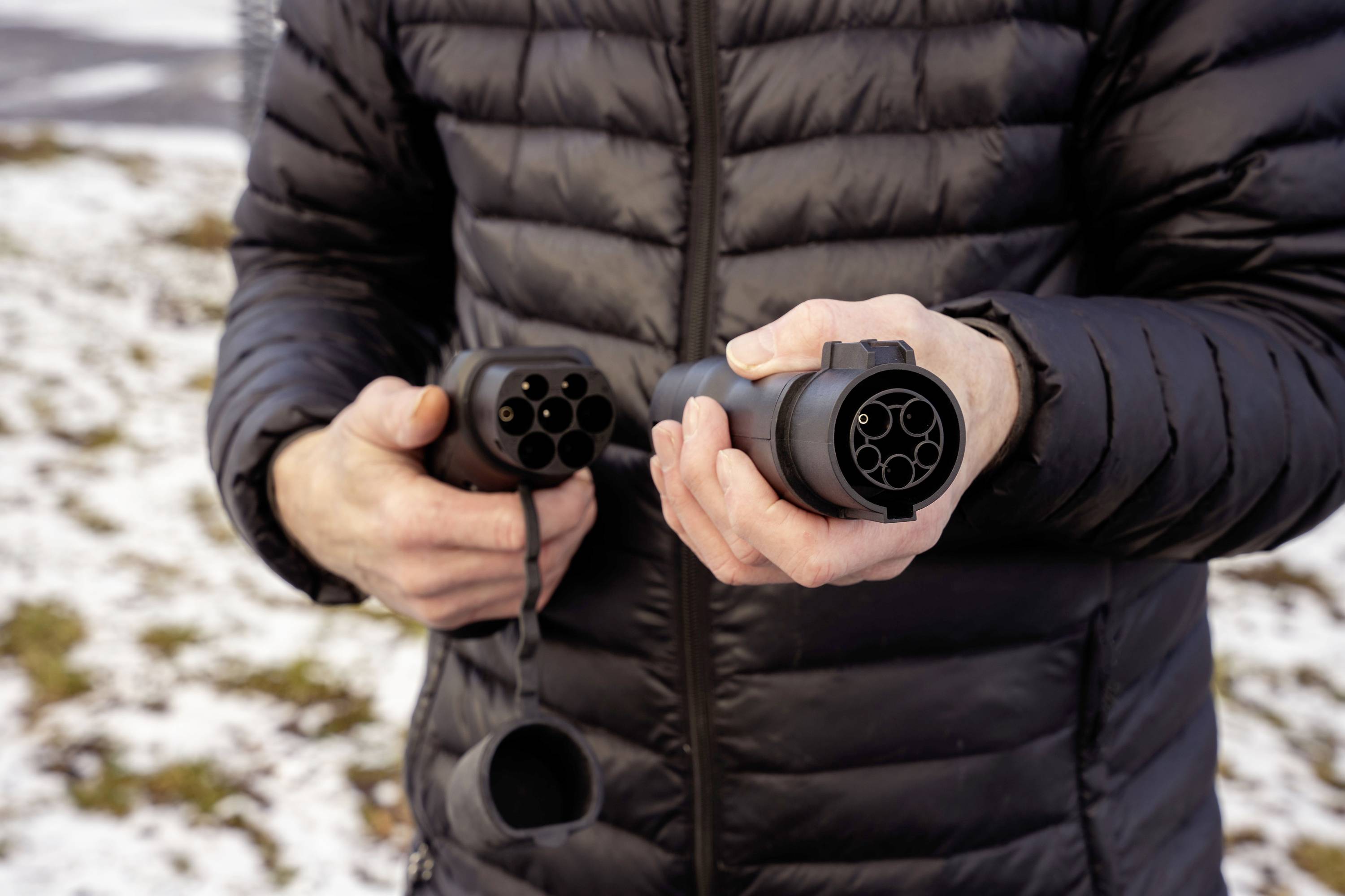A person is holding an electric charging cable in both hands. The background shows snow-covered ground.