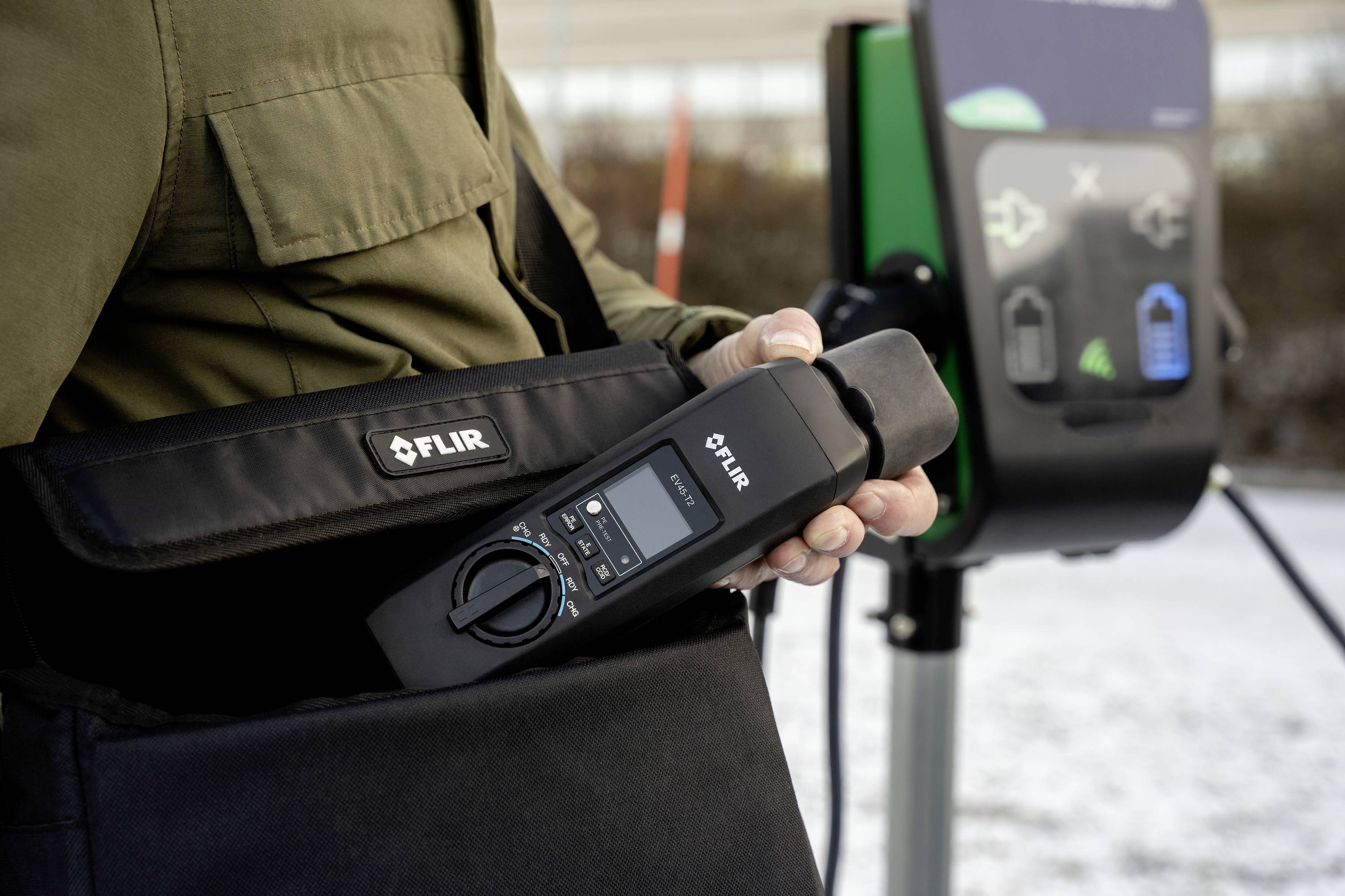 A person is holding a FLIR measuring device that they are pulling out of a bag. In the background, there is an electric vehicle charging station.