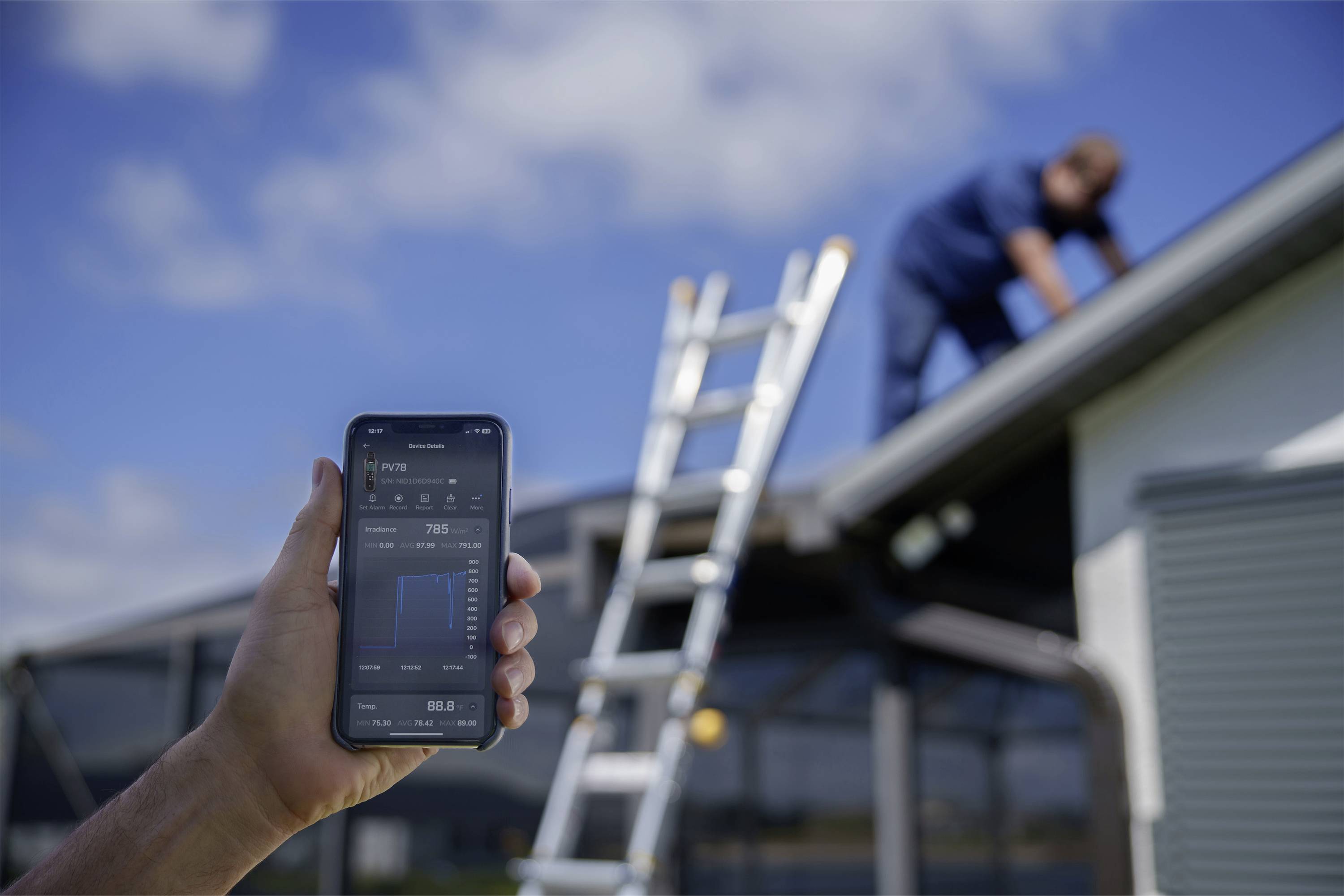 A smartphone displays solar panel data, while a person works on a solar installation on a roof in the background.