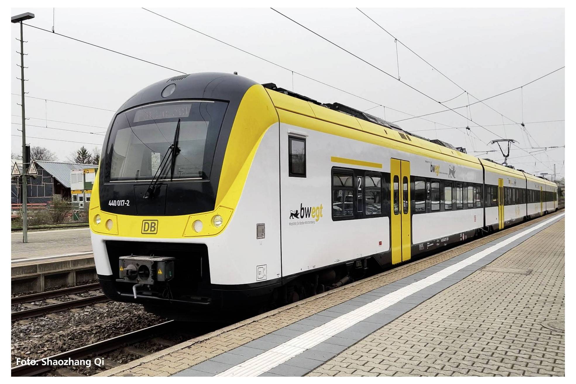 A yellow regional train with the inscription 'bwegt' is standing on the platform. Railway tracks and buildings can be seen in the background.