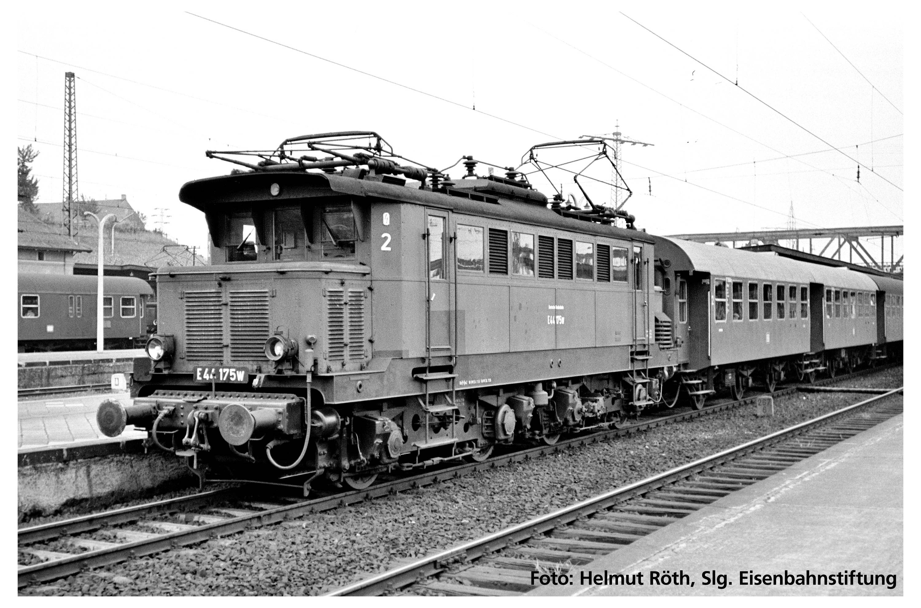A historic electric locomotive with attached carriages stands on a railway track at a station. Text: 'E 44 - 175W'.