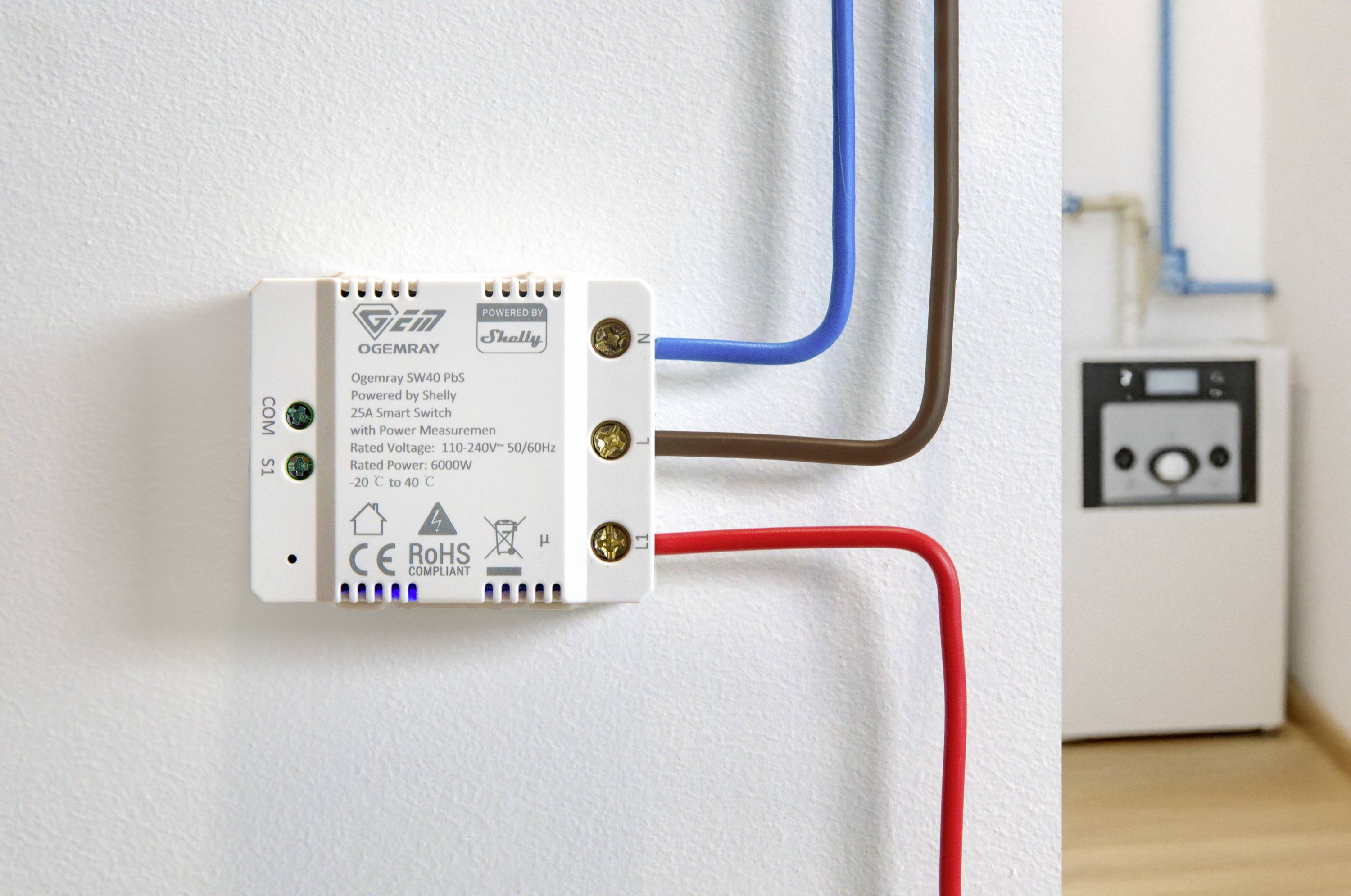 An electrical switch with blue, brown, and red wires is mounted on a white wall. A boiler can be seen in the background.