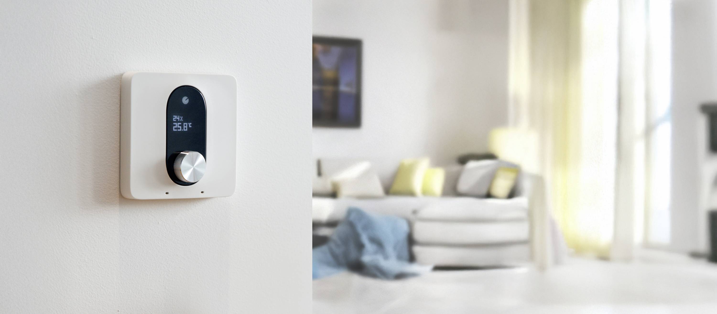 A modern thermostat on a white wall shows 26 degrees Celsius. In the background, a bright living room with a sofa and cushions.