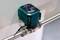 A green water meter sensor is attached to a horizontal pipe. The sensor measures water consumption digitally.