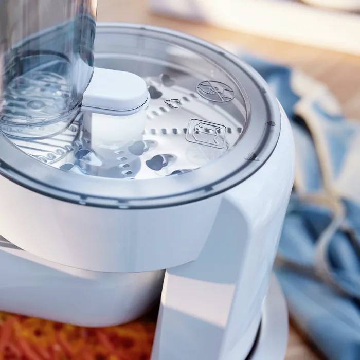 A white kitchen mixer with a transparent lid on a wooden table. Kitchen equipment blurred in the background, with a blue cloth.