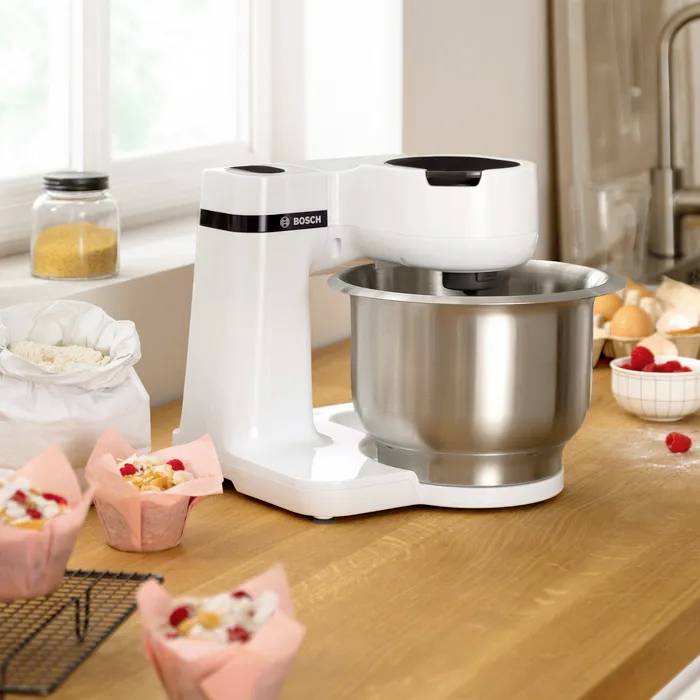 Mixer on a wooden table, surrounded by ingredients such as flour, eggs and berries. Finished muffins in pink cases are ready.