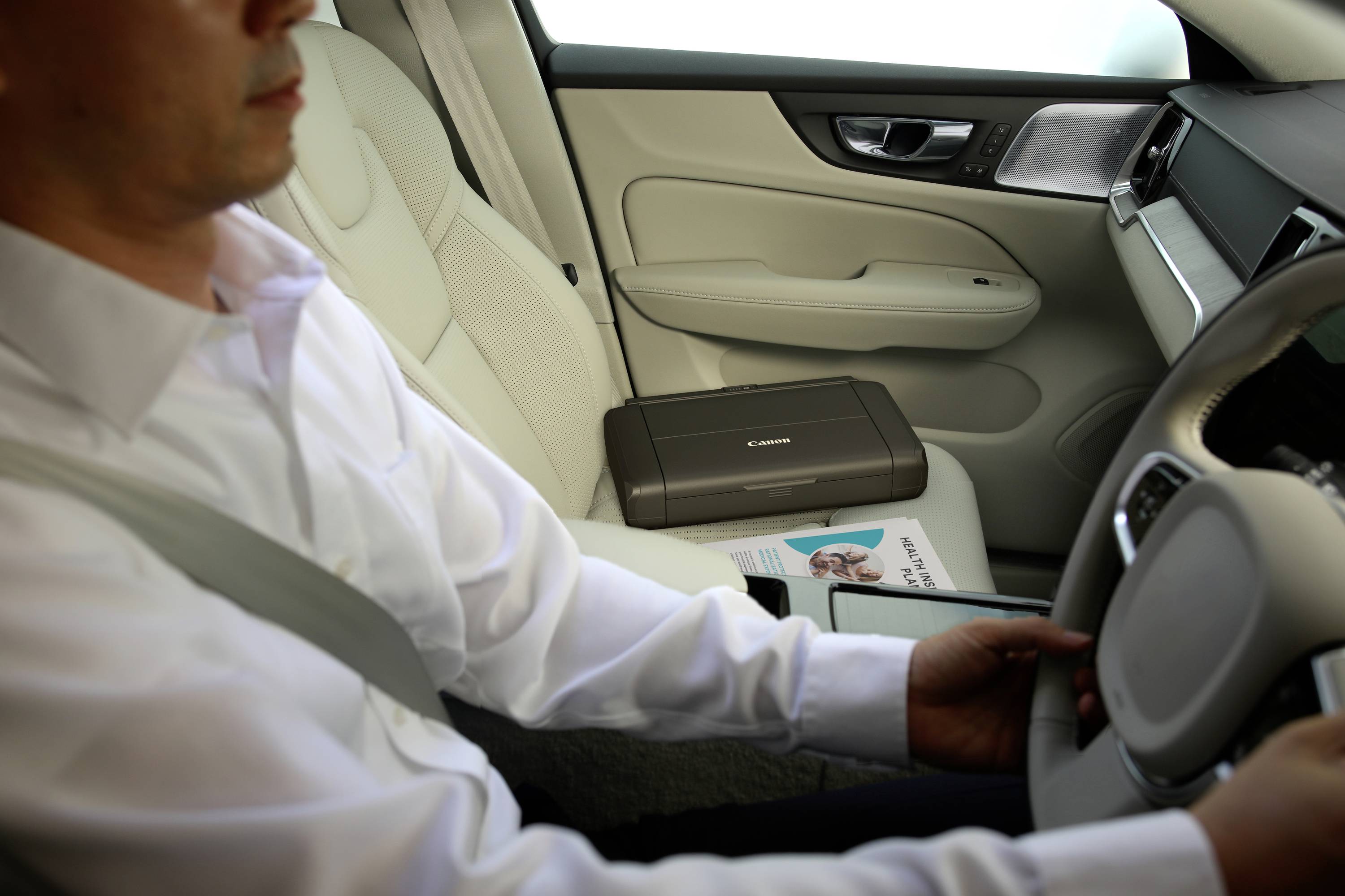 A person is driving a car, holding the steering wheel. A portable printer and magazines are lying on the passenger seat.