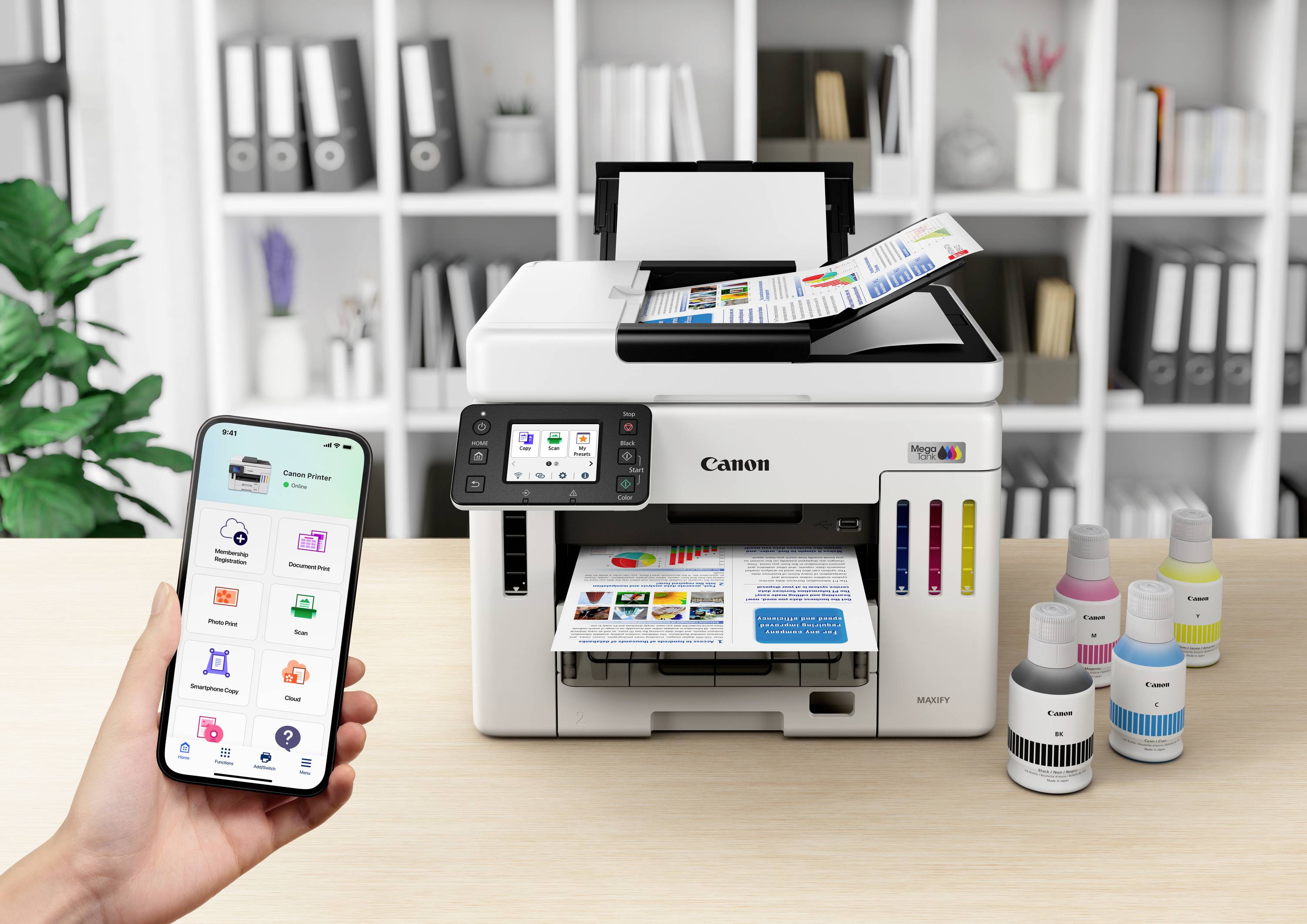 A person is operating a printer via smartphone. The printer on a table is printing colour documents. Ink bottles are standing to the right beside it.