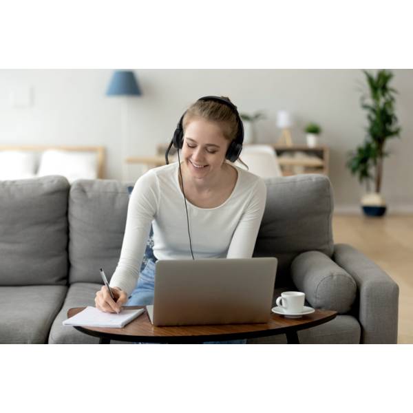 A woman is sitting on a sofa, wearing headphones and taking notes while looking at a laptop. A cosy living room setting is visible in the background.