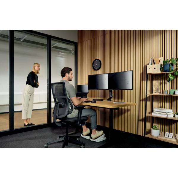 A man is working at a desk with two monitors. A woman is standing in the background. The room appears modern and tidy.
