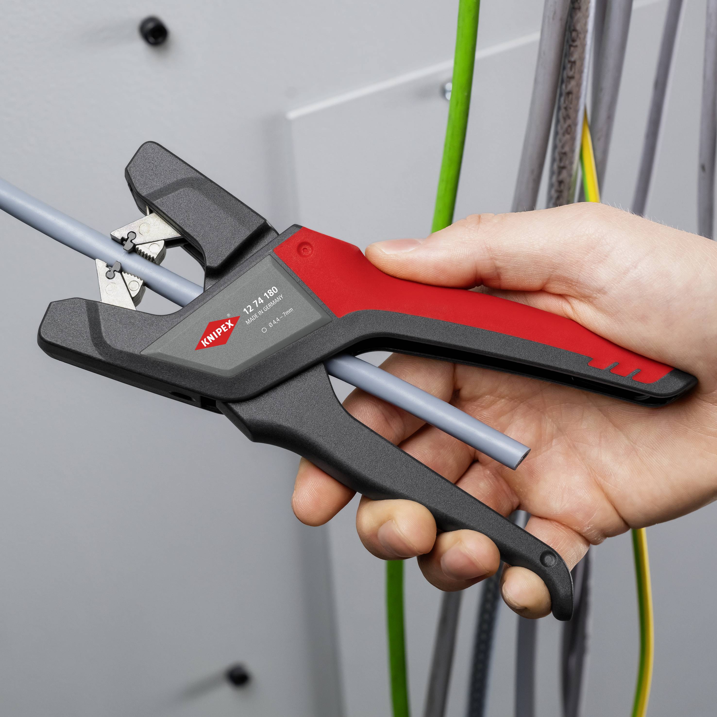 A hand is holding a red and black side cutter and cutting a grey cable. Other cables are visible in the background.