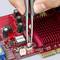 A hand is using tweezers to precisely position a small metal pin on a red computer circuit board.