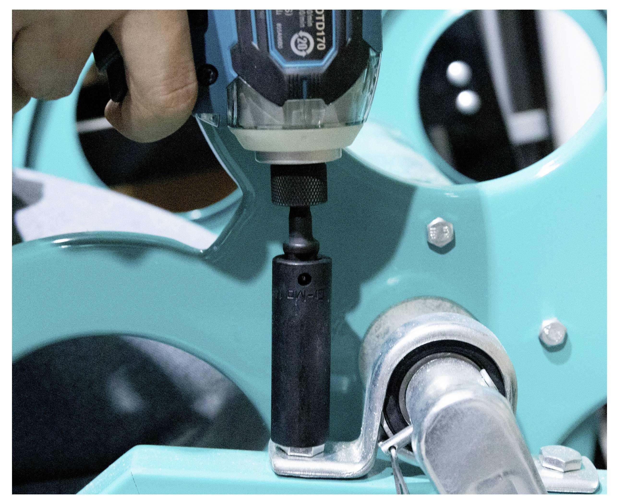 A person uses a power drill with a socket attachment to adjust a metal component of turquoise machinery, tightening a bolt.