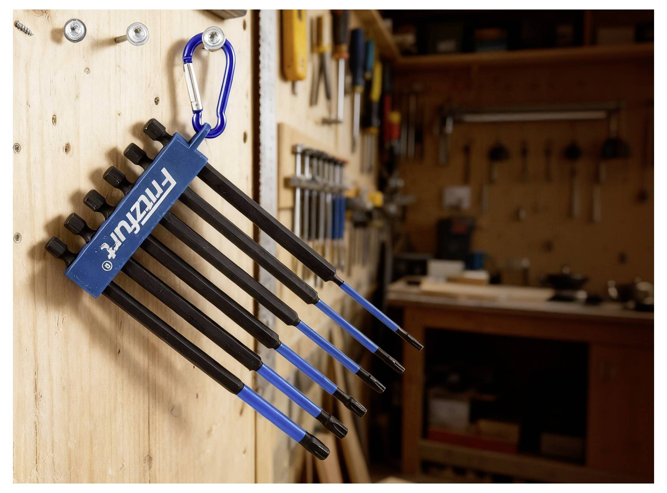 A set of hex keys hanging on a workshop wall, attached with a blue carabiner. Various tools and workbenches are visible in the background.