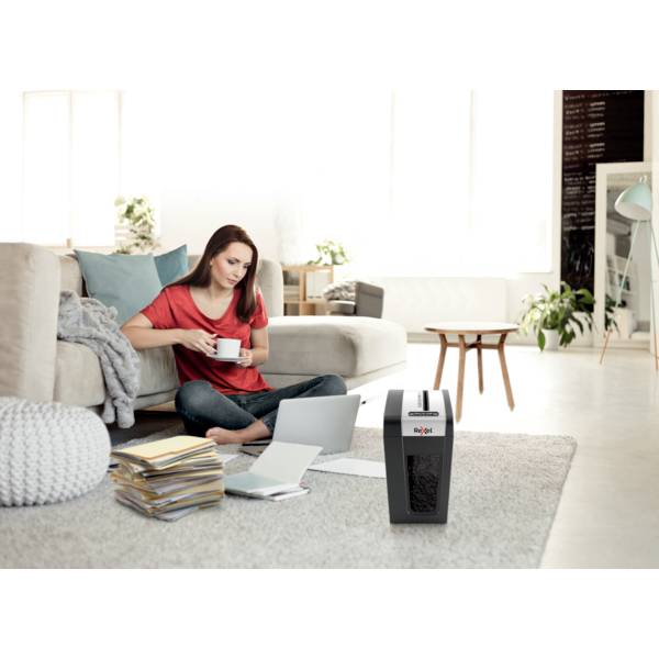 A woman is sitting on the floor of a living room, reading documents and drinking coffee. A paper shredder is next to her.