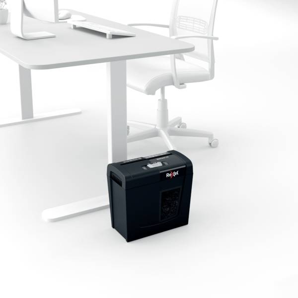 A black paper shredder sits beneath a white office desk next to a white office chair. In the background, a computer and keyboard can be seen on the desk.