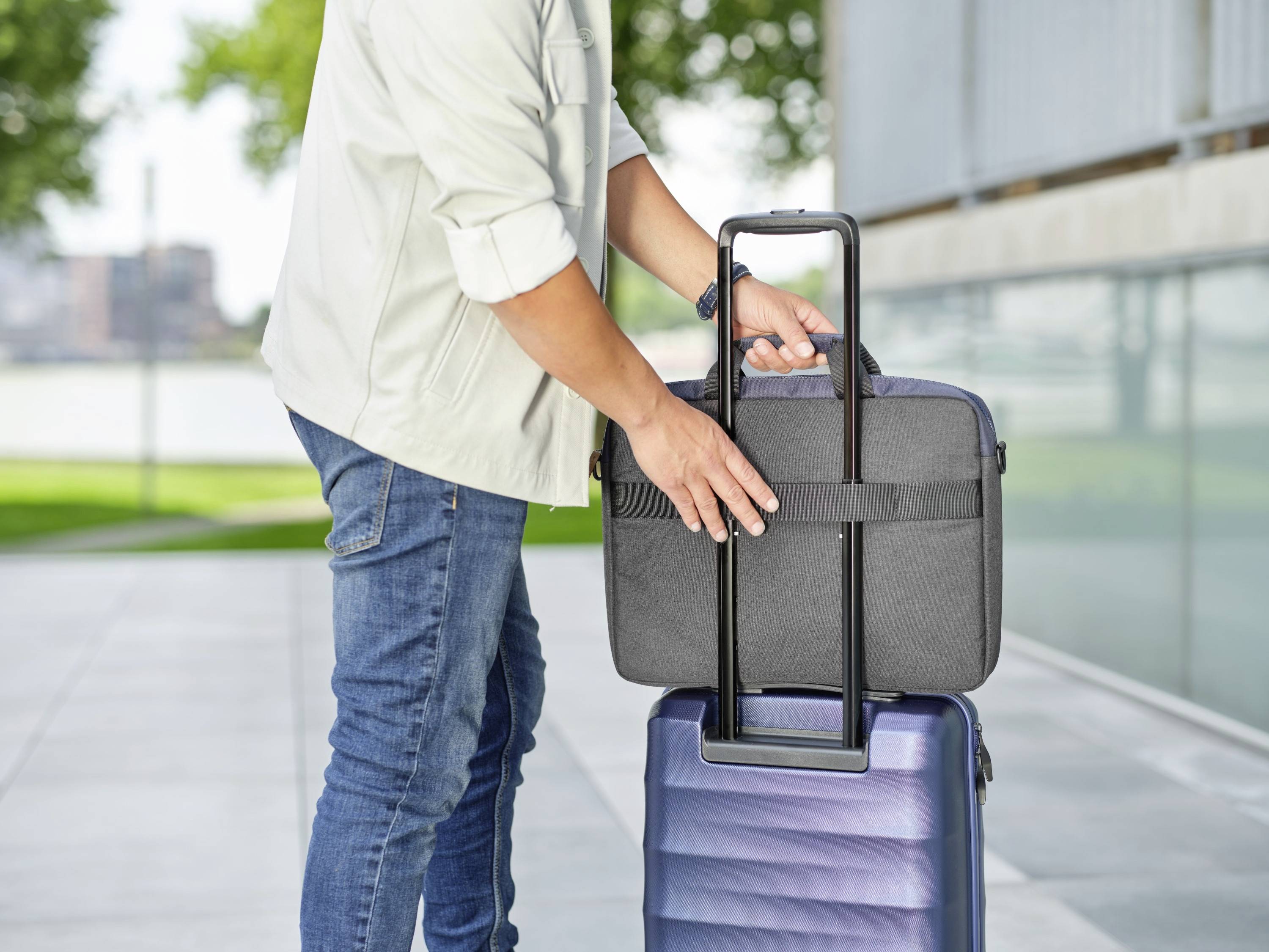 A person is attaching a laptop bag to the handle of a blue rolling suitcase outdoors.