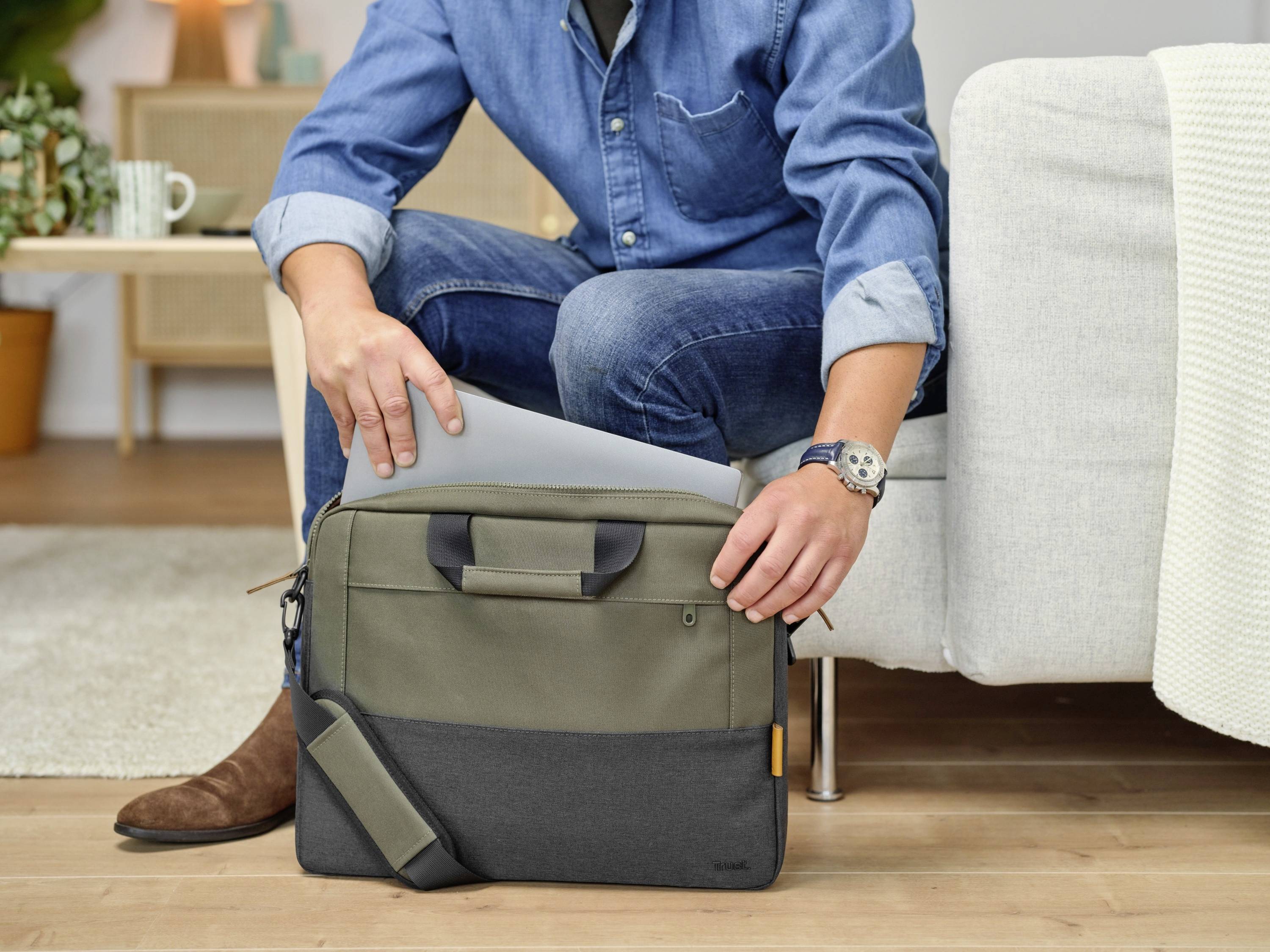 A person in a denim shirt is placing a laptop into a two-tone briefcase. Furniture and plants are visible in the background.