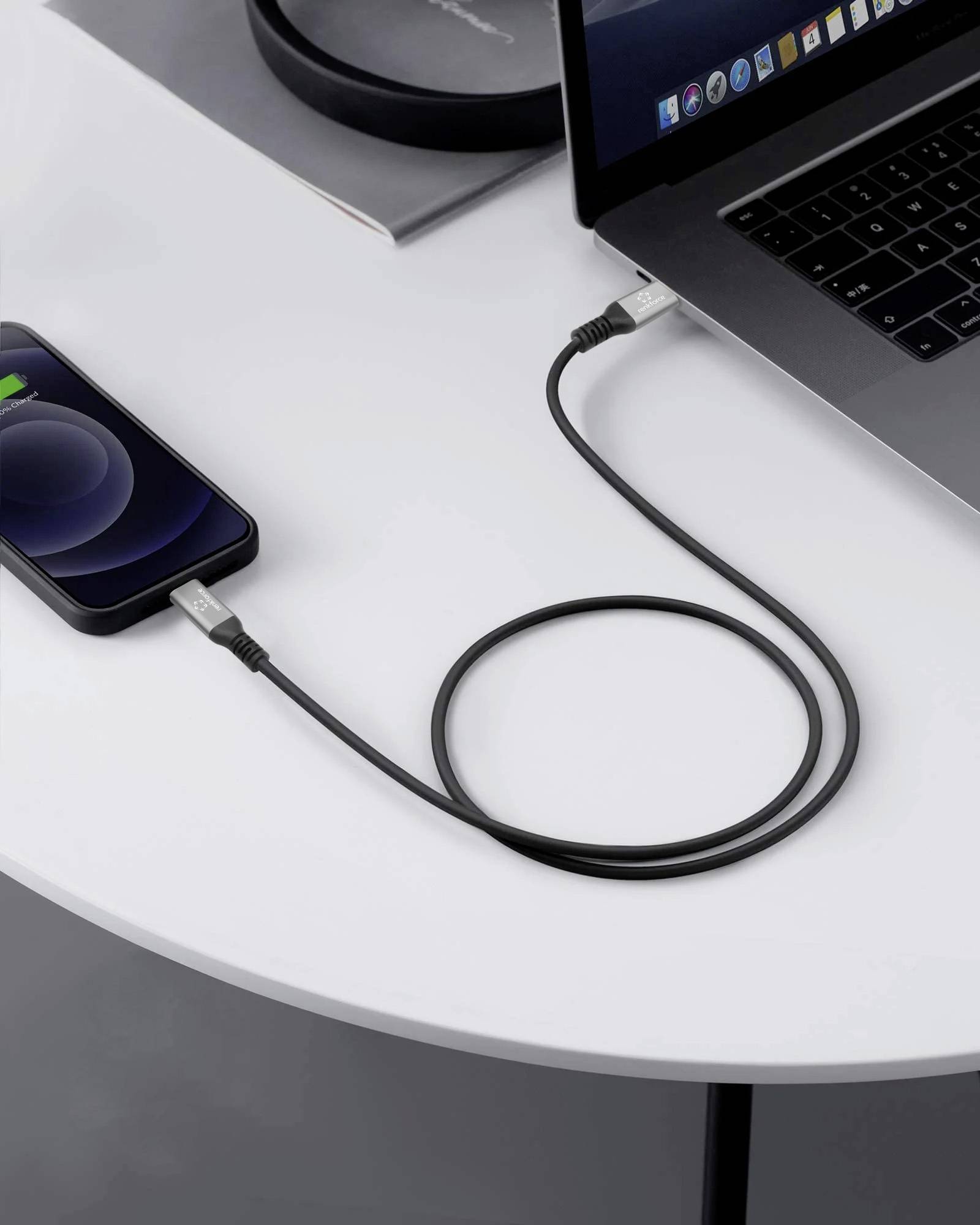 Smartphone charging via a cable connected to a laptop that is sitting on a white table. The phone displays a charging icon.