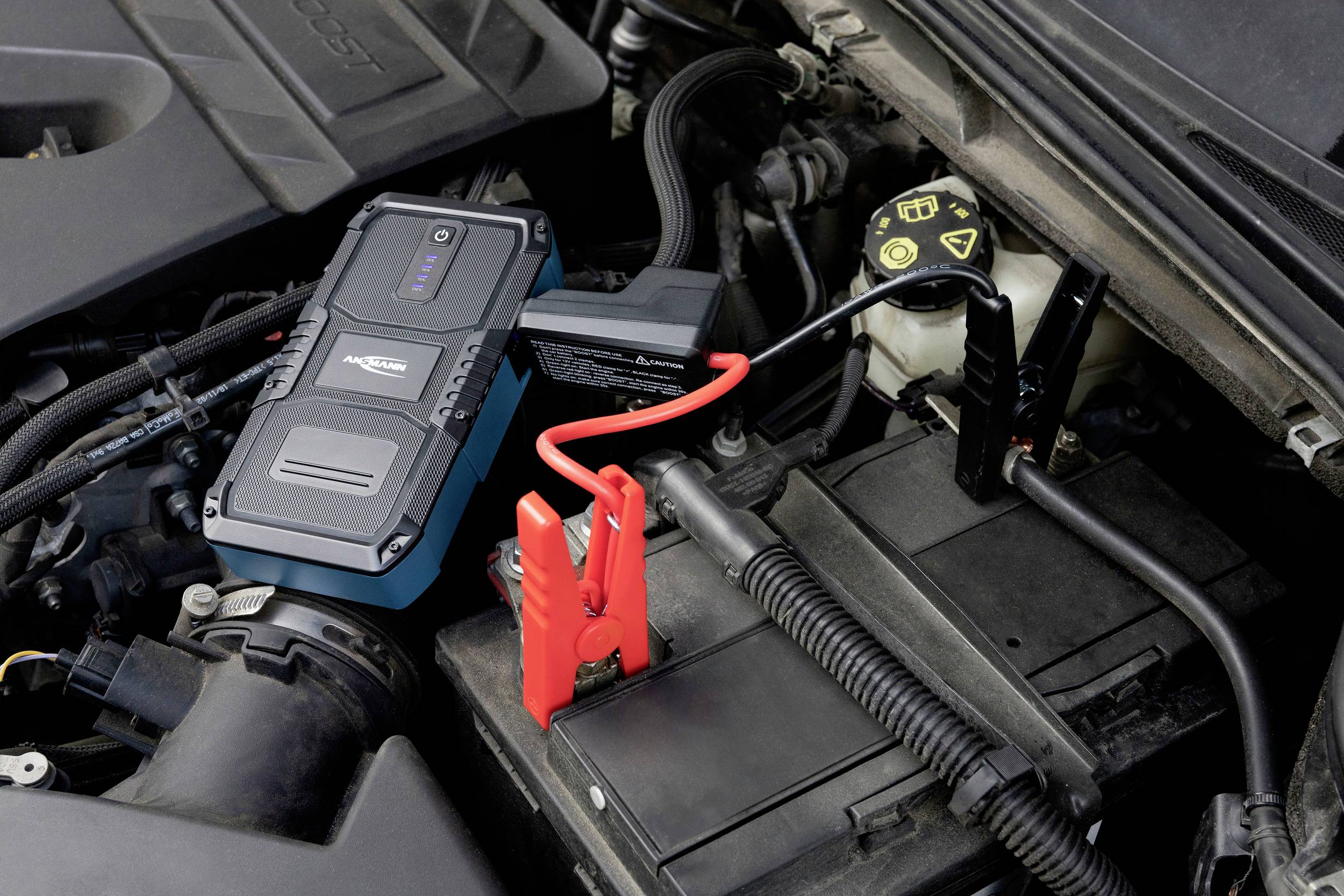 A jump starter is connected to a car battery. A red jump lead connects the device to the positive terminal of the battery.
