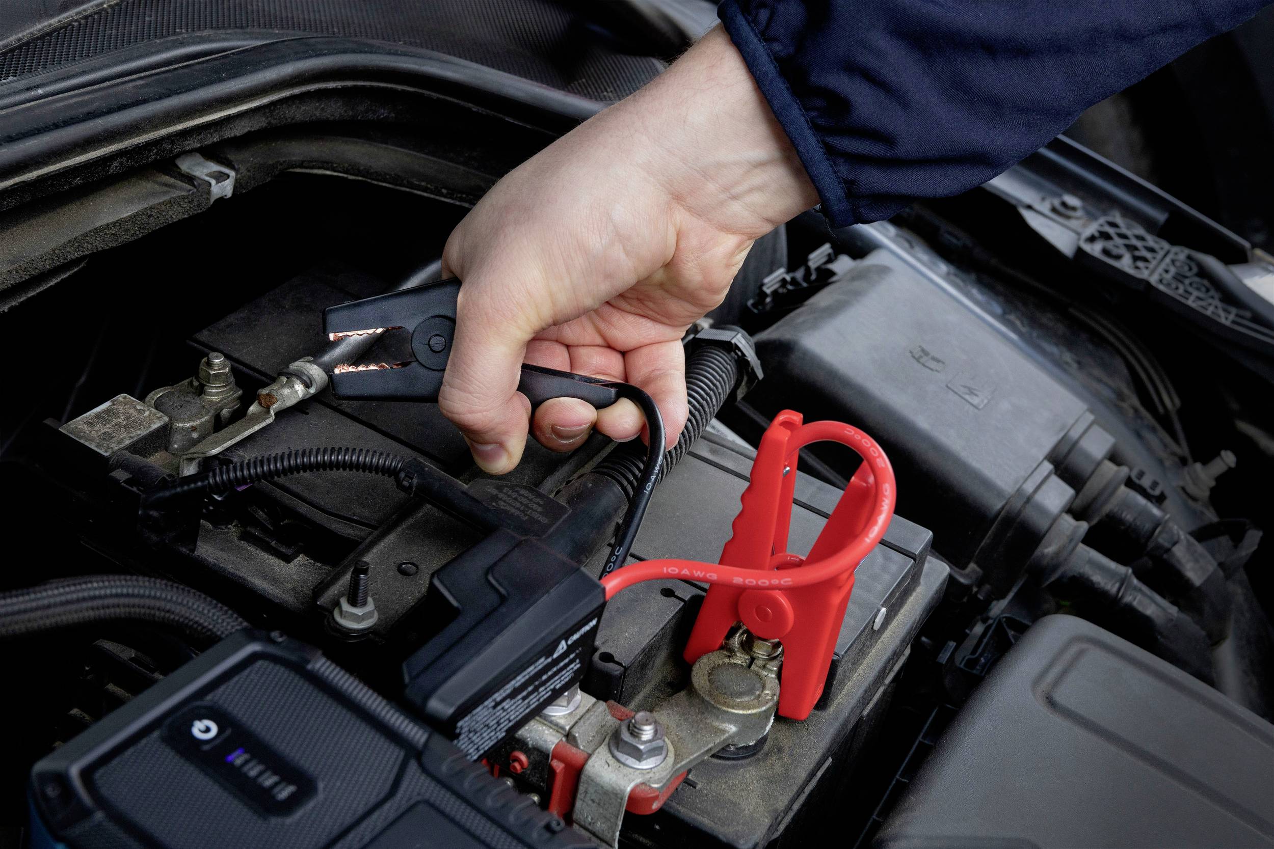 A hand connects a black jump lead to a car battery to charge the vehicle battery.