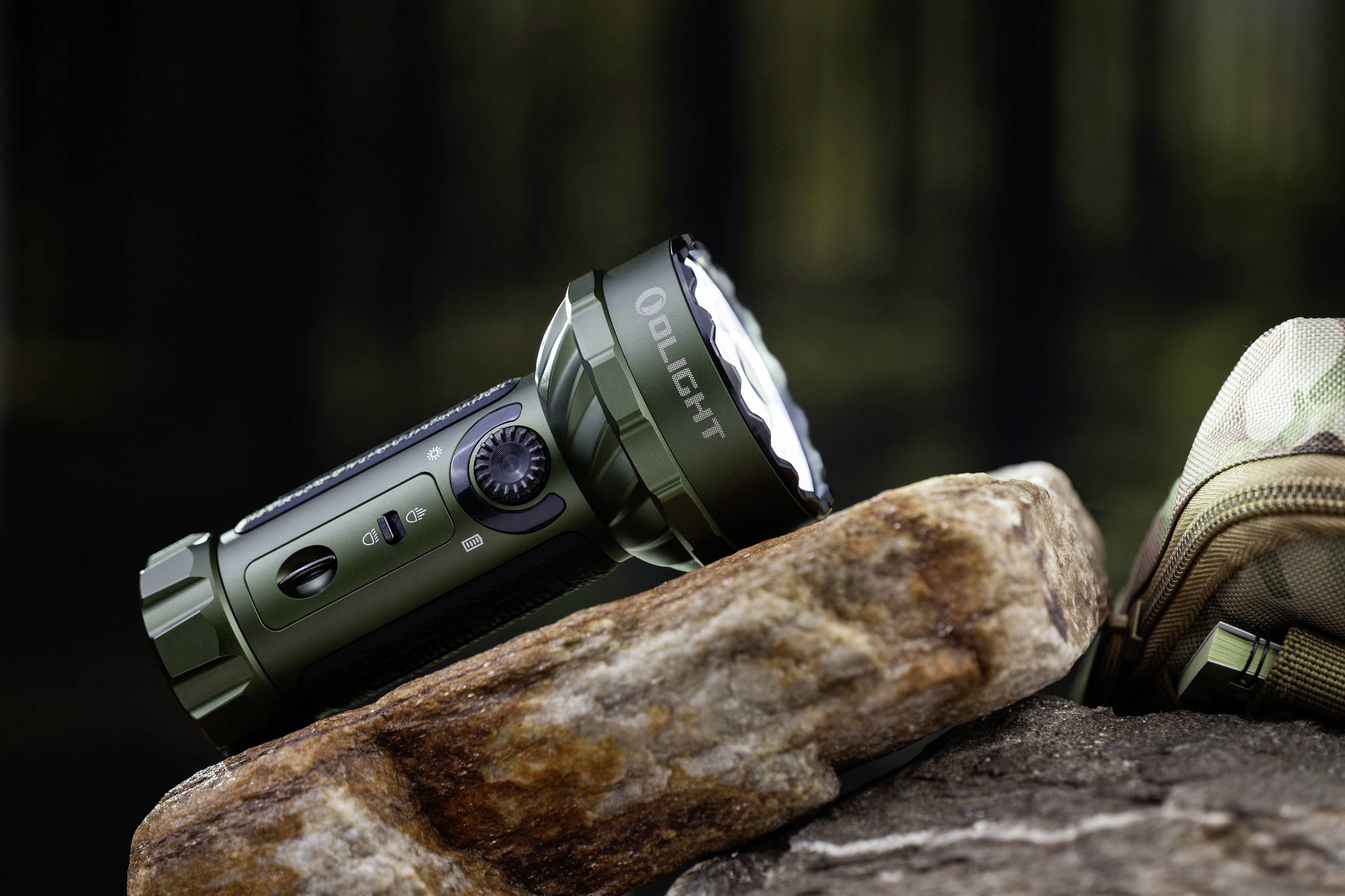 An olive green torch lies diagonally on a rock in the woodland. A partially visible camouflage rucksack is positioned on the right side of the image.