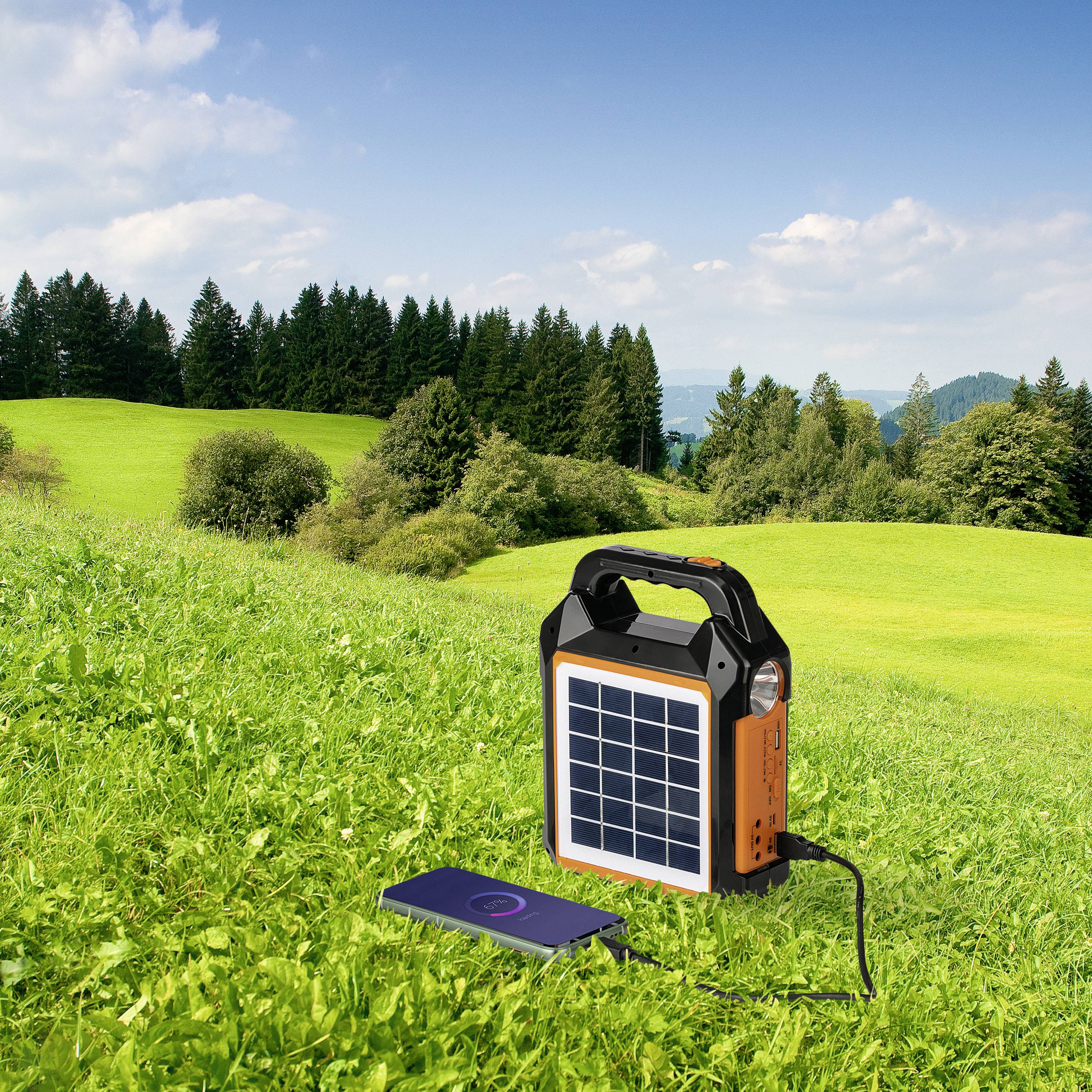 A portable solar panel charger is charging a smartphone on a green meadow. Trees and a blue sky are visible in the background.