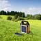 A portable solar panel charger is charging a smartphone on a green meadow. Trees and a blue sky are visible in the background.