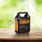 A portable, orange and black solar radio compact device on a wooden table. Musical notes indicate it is playing music. Background green and blurred.