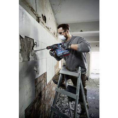 A man is wearing safety glasses and a respiratory mask, using an electric drill to remove tiles from a wall, standing on a ladder.