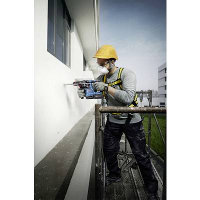 A construction worker wearing a hard hat and mask is drilling into a white wall with a drill, standing on scaffolding.