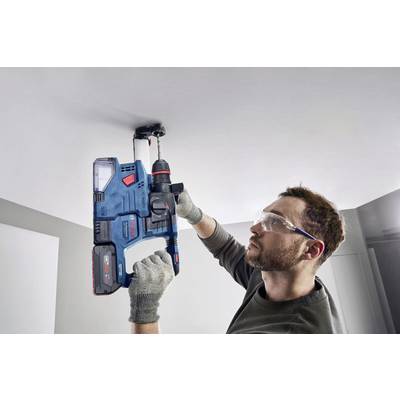 A person is drilling into a ceiling with a blue hammer drill. They are wearing safety glasses and gloves.